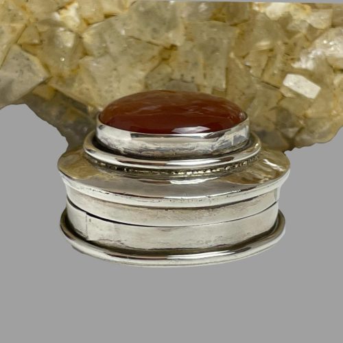Main photo of silver & carnelian pill box seen from an eye level. The box is displayed in front of a rock of river quartz which is has a pale yellow crystal hue. This is reflecting on the silver area of the pill box lid. As with a lot of pill boxes with a stone set in the lid, this one has more depth to the lid than the base.