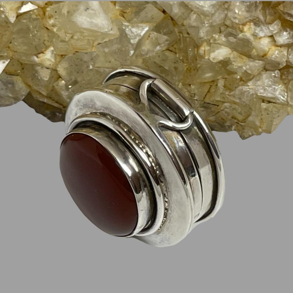 Photo of silver & carnelian pill box displayed on its side with the carnelian at top of lid facing the bottom left corner of photo. The solid sturdy hinge is visible in this shot.
