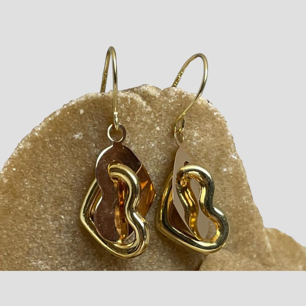 Close up photo of 14kt gold heart earrings still displayed hanging from some natural desert sand rose. The rose gold solid flat hearts are clearly visible and one side of the open yellow gold hearts pierces through with the remaining side of the heart hanging below the rose gold one. There is no hallmark on the hearts but it is possible to make out the hallmark on the hook clasp.