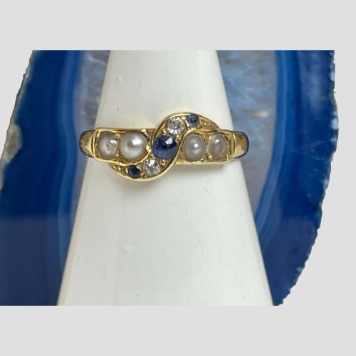 Main photo of Victorian sapphire diamond ring displayed forward facing on a cone shaped stand with a slice of blue agate in the background. The main band of the ring houses the seed pearls with an S shaped band crossing the centre with the largest of the sapphires at centre. On either side of the central sapphire are the diamonds followed by smaller sapphires at either end. An unusual but effectively striking setting. Beautiful blue colour on the sapphires.