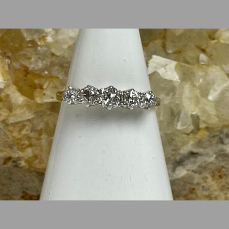 Main photo of 18kt gold diamond ring shown displayed on a cone shaped stand with the ring front forward facing. The 5 diamonds cover nearly all of the stand with just a small area of band visible at either end. The diamond to the centre is the larger of the 5 with the outer 2 graduating down in size. There is a rock of natural river quartz in the background.