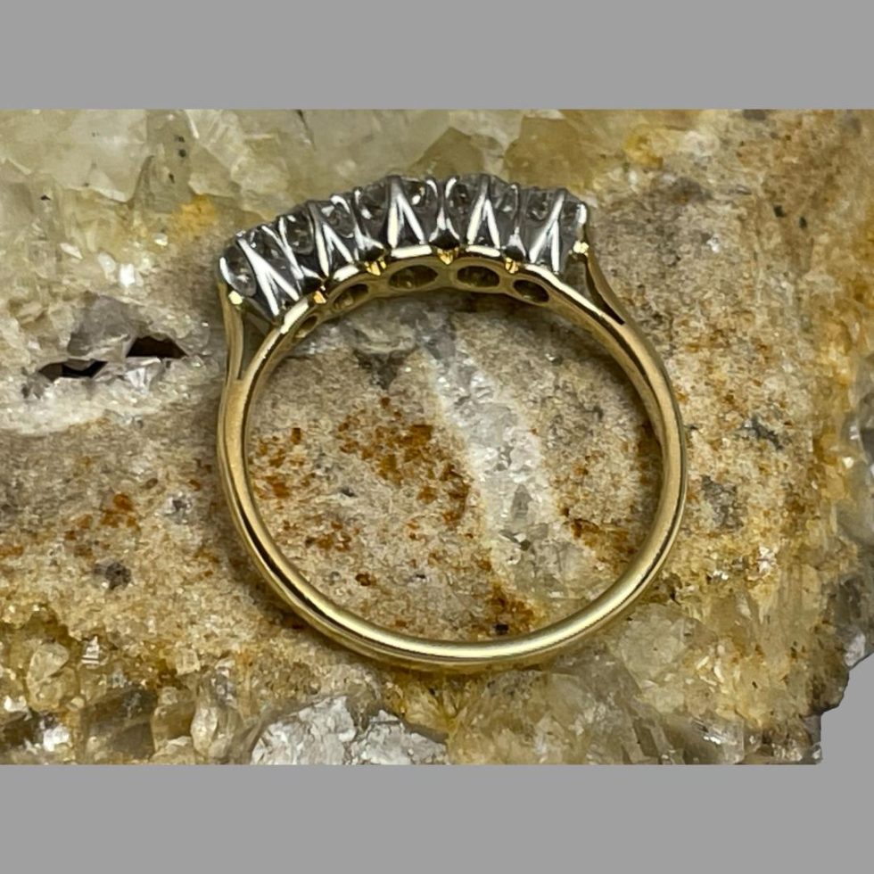 Photo of 18kt gold diamond ring displayed on top of some natural river quartz. We are looking straight down from above at it. The front diamond end of the ring is facing off centre to the top. The ring is still on top of some natural river quartz.