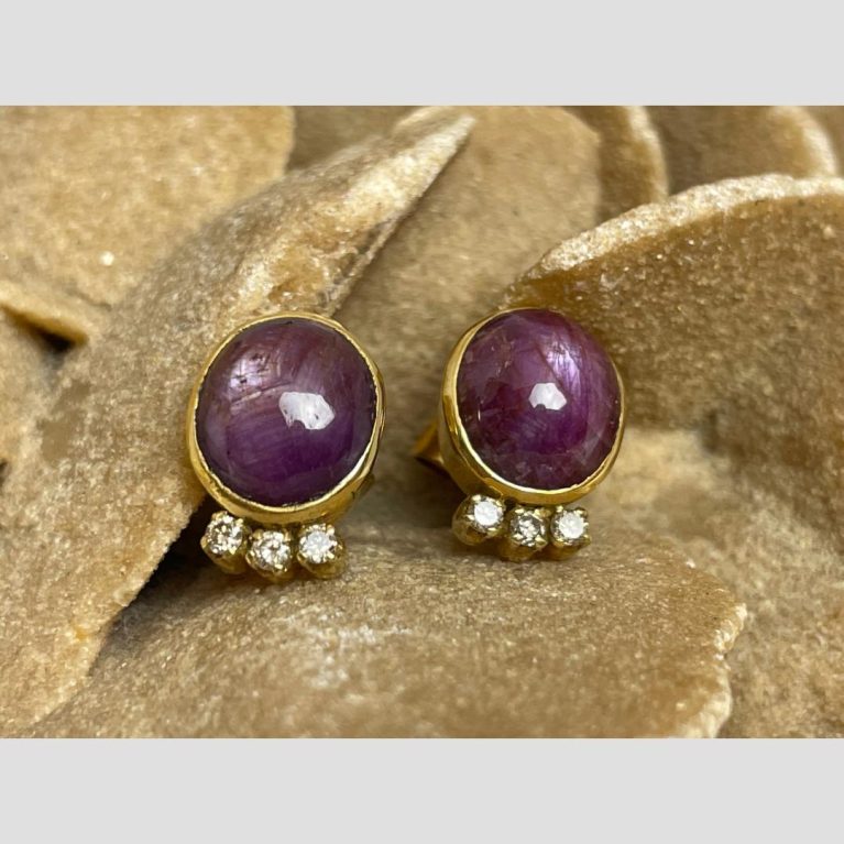 Main photo of star ruby and diamond earrings displayed on top of a rock of natural desert sand rose. The stones are forward and slightly upward facing with the stars within showing alongside the studio lighting. The 3 diamonds are in a row below the ruby and sparkle beautifully.