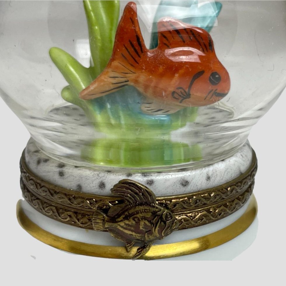 A close up of the goldfish in the bowl and the fish on the opening clasp. The top of the box below the bowl is white with black dots like a dalmation.