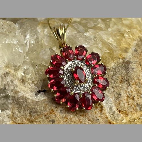 Main photo of gold ruby diamond necklace focusing just on the ruby and diamond pendant & bail. The pendant is laid on a rock of natural river quartz with the light shining on it enhancing the beautiful red of the rubies.