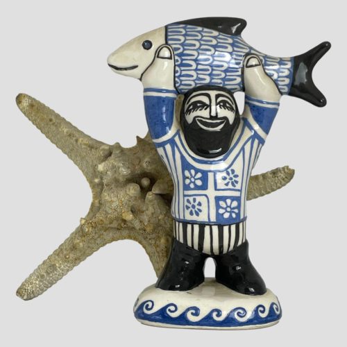 Main photo of fisherman by Bernard Moss seen from an eye level. The fisherman is holding both arms up and holding a fish above his head. He has a face of absolute joy donning a cheeky big smile . Like his female companion, the colours are cream, Cornish blue and black. The fish has black fin and tail with a smiling black mouth and blue eyes with black rim. The fisherman has black features including a beard and moustache and is wearing black boots. His trousers are black and cream vertical striped and look like briefs as boots are high. He has quite a fancy jumper on in blue and cream with 4 panels with flowers in a panel at front, cream and blue striped upper arms and plain blue lower arms. He stands on a cream base with wavy pattern all around the sides. A starfish is displayed behind him adding to the seaside theme.