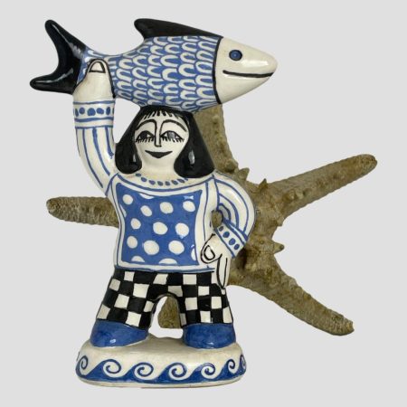 Main photo of Bernard Moss fisherwoman figure seen from an eye level with the figure facing forward. She is a fabulous fun figure with her left arm on her hip and the other holding a large fish aloft by its tail above and resting on her head. The colours are uniform and simple - just cream, Cornish blue and black and the patterns of her clothing and the scales of fish make this piece strikingly cheerful and a sight to behold. Both the fish and woman have a cream base colour. The fish has black fin, tail, eye & smiling mouth and the scales are blue as is the eye ball. The woman is wearing black and cream chequered trousers, blue shoes, blue and cream dotted top with cream long sleeves with blue stripes. Her hair and facial features are all in black and she is donning a wide smile. She stands on a base of cream with blue wave pattern around the sides. There is a starfish in the background to enhance the seaside theme although in this case, it was probably unnecessary!