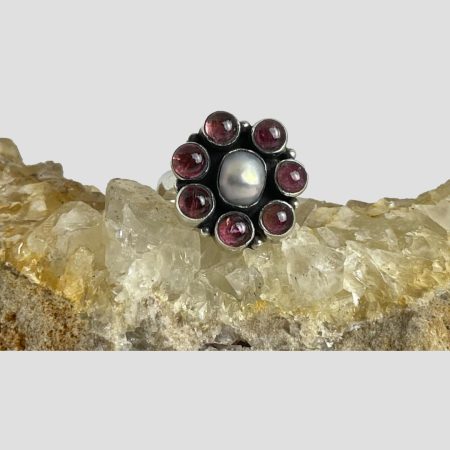 Main photo of silver garnet pearl ring displayed on top of a rock of natural river quartz with the ring front forward facing. The ring has an oval shaped pearl to the centre with the 7 garnet cabochons laid out like flower petals. Very simple design but very effective and also quite festive!