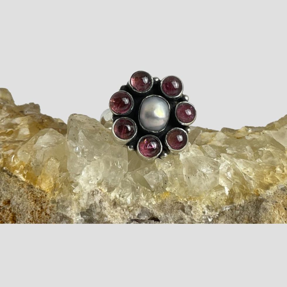Main photo of silver garnet pearl ring displayed on top of a rock of natural river quartz with the ring front forward facing. The ring has an oval shaped pearl to the centre with the 7 garnet cabochons laid out like flower petals. Very simple design but very effective and also quite festive!