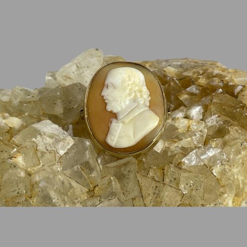 Main photo of 19th century Shakespeare cameo displayed on top of a rock of natural river quartz with the brooch front forward facing. The Bard of Avon is profiled facing left and is expertly carved in great detail. The shell background colour is a medium burnt orange with Shakespeare splendid in cream and white.