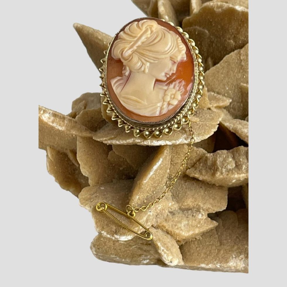 Main photo of 9 karat gold cameo brooch displayed balanced on a rock of natural desert sand rose with the brooch front forward facing. The lady in profile has her head on the incline so facing down to the lower right. The safety chain and pin are extending down below the brooch.
