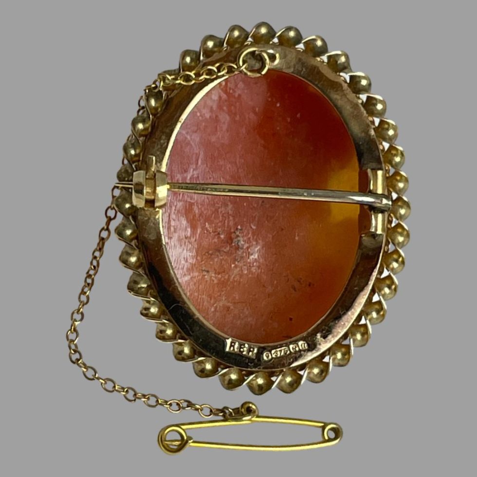 Photo of the back of the 9 karat gold cameo brooch displayed upright with the safety chain and pin cascading down to the left and the pin resting in the foreground. The hallmark is just visible along the bottom rim at the back.