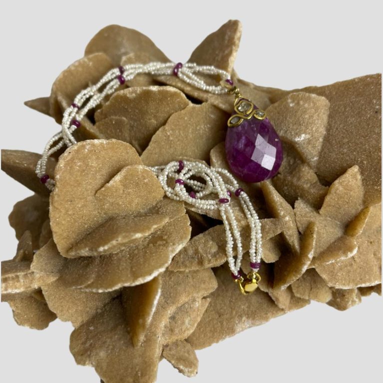 Main photo of ruby diamond pearl necklace displayed on top of a rock of natural desert sand rose. The ruby pendant is displayed with the diamonds side on display and the seed pearl and ruby bead necklace is draped over the sand rose with the gold clasp dangling in the foreground just below the pendant.