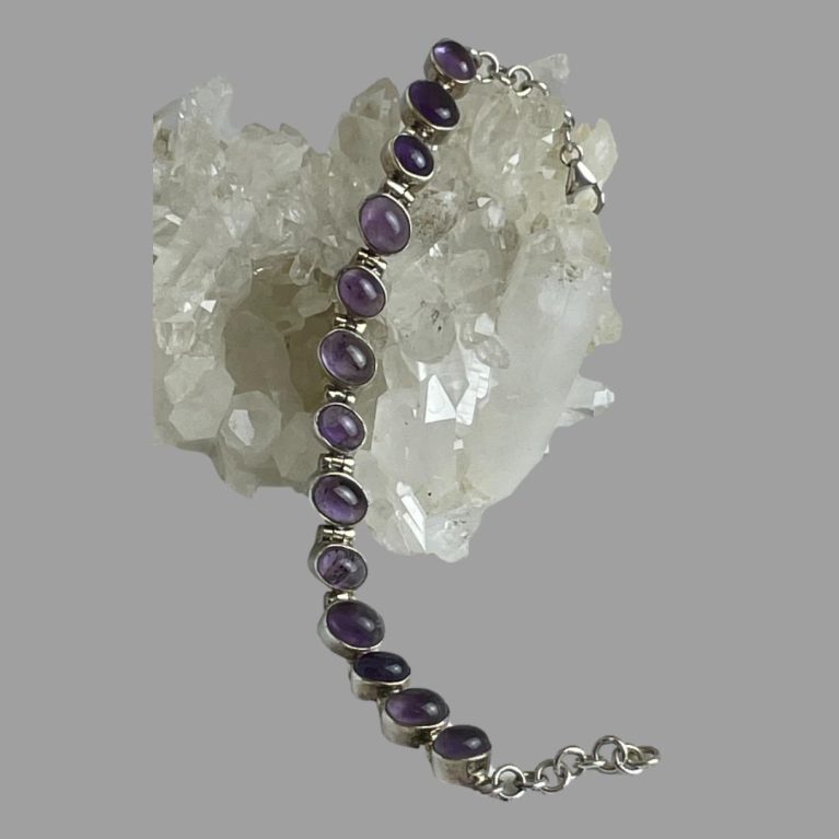 Main photo of sterling silver amethyst bracelet shown unclasped and laid across a rock of natural quartz crystal. Not easy to see when it is unclasped but this bracelet has an interesting and unusual design where the links are set at an angle so the amethyst stones appear to be leaning. Also when the clasp is closed, the bracelet sets rigid so appears like a bangle rather than a bracelet.