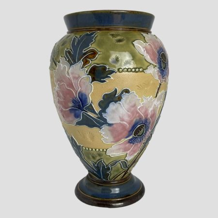 Main photo of Royal Doulton poppy vase seen from an eye level and with the decorated side on display. The vase has a dark earthen blue rim to the top and base, a peachy yellow centre and an earthy olive green below the rim at top and above the rim at base. The poppy decoration which covers only half of the vase have the same blue for the tall leaves and a lighter blue at the centre of the poppies. The poppy petals are pink in the centre and a very pale pink to their outer edges. The combination of these colours work extremely well and appear to highlight each part of this wonderful collectable vase.