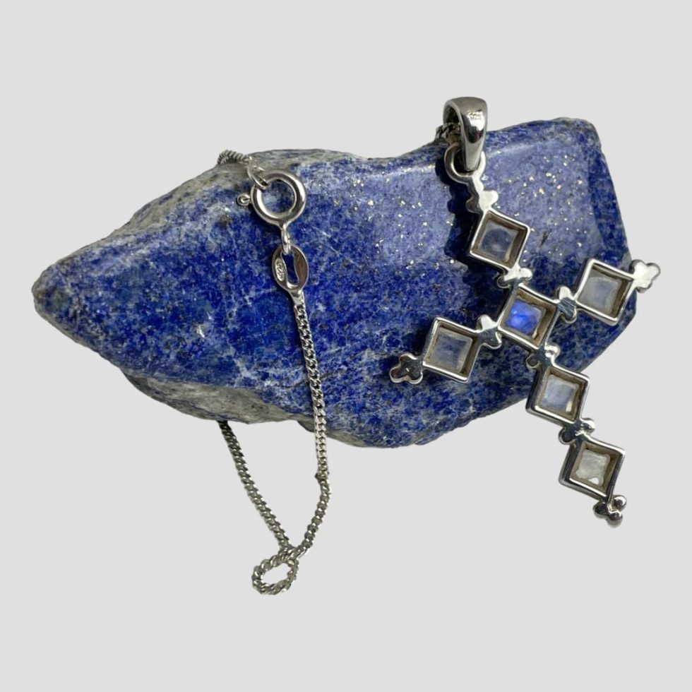 Photo of the back of the silver moonstone cross &chain showing the open back so the stones are still visible. The clasp area of the chain is hooked around the end of the lapis to the left of cross.