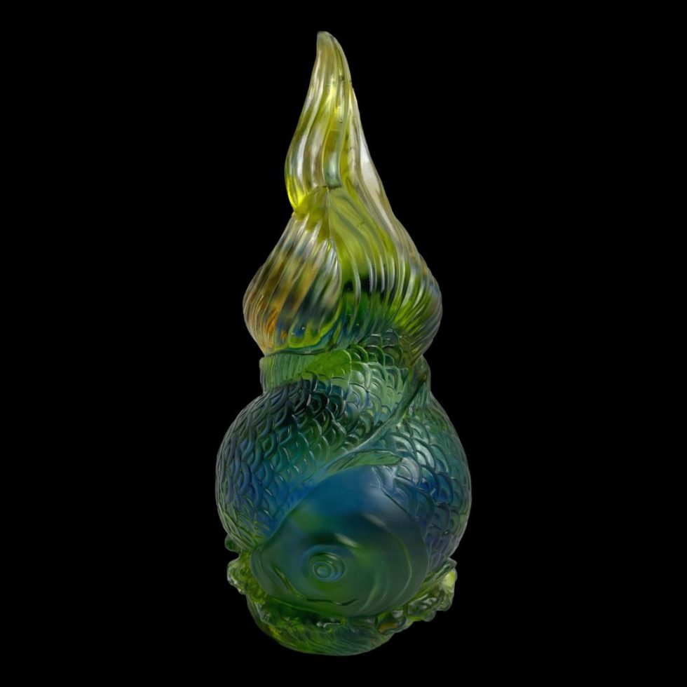 Main photo of Liuli Gongfang fish sculpture seen from an eye level with one of the fish heads in the lower foreground. The piece is almost like a flame shape with the delicate tail fins in a light green yellow at the top like tip of flames. The colours deepen as we go further down to green mixed with blue finishing with a darker green at the base. The mix of these colours give movement to the piece as though we are swimming with the fish. A beautiful piece of art glass displayed against a black background enhancing the colours all the more.