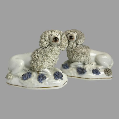 Main photo of late Georgian Staffordshire poodles displayed again with one slightly in the foreground. The head ends of both dogs are in the centre of photo and they are both facing towards the camera but looking slightly upward with their eyes. There are 3 blue tufts on the base alongside their bodies which can only be flowers. The poodles tail is also styled in same manner as mane.