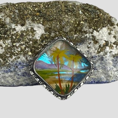 Main photo of silver butterfly wing brooch shown leaning against a natural rock containing lapis . The brooch is shown in diamond position as opposed to square as the image works within the diamond. There are 2 palm trees in the foreground with yellow palm fronds and a reddish brown trunk. Behind the trees is a beach scene with mountains in the distance the beautiful turquoise flashes of the butterfly wings bring this scene to life.