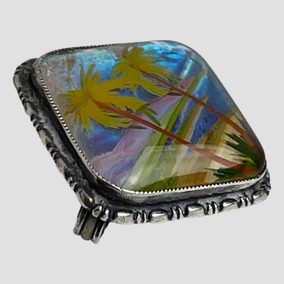 Photo of silver butterfly wing brooch laid on a flat surface and seen from a side angle. The top of the palm tress are to the top left of the photo.
