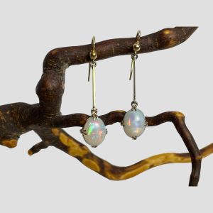 Main photo of antique gold opal earrings displayed hanging on natural wooden twig with the earring front forward facing. The fasteners are hooks from which a delicate gold bar hangs down which in turn has an opal cabochon attached at the end.