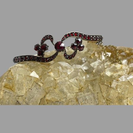 Main photo of antique garnet bracelet shown displayed on top of a rock of natural river quarts with the bow like front of the bracelet facing forward. The main band of the bracelet finishes at the front in a upward and downward curve with the large central garnet as the centrepiece. Opposite to the main band are short extensions with 3 garnets at the end of each giving a feel of the ends of a bow. There are garnets all the way round this bracelet right up to the clasp. The gold safety chain can be seen extended out on the right. There is a lovely festive quality to this bracelet.