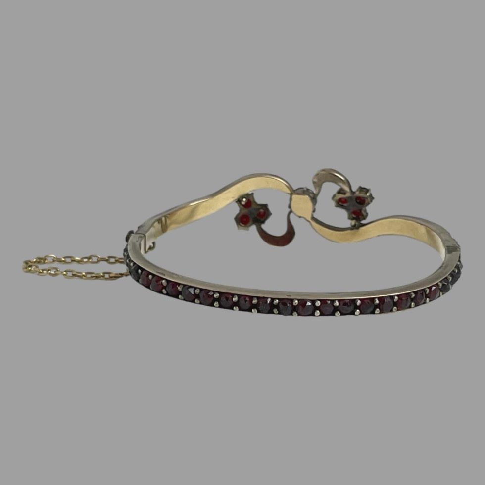 Photo of the back of antique garnet bracelet displayed on a flat surface and seen with the bracelet back in foreground. The garnet setting in the bow like area shows that the backs are all covered except for the back of the 3 garnets at the end of the bow.