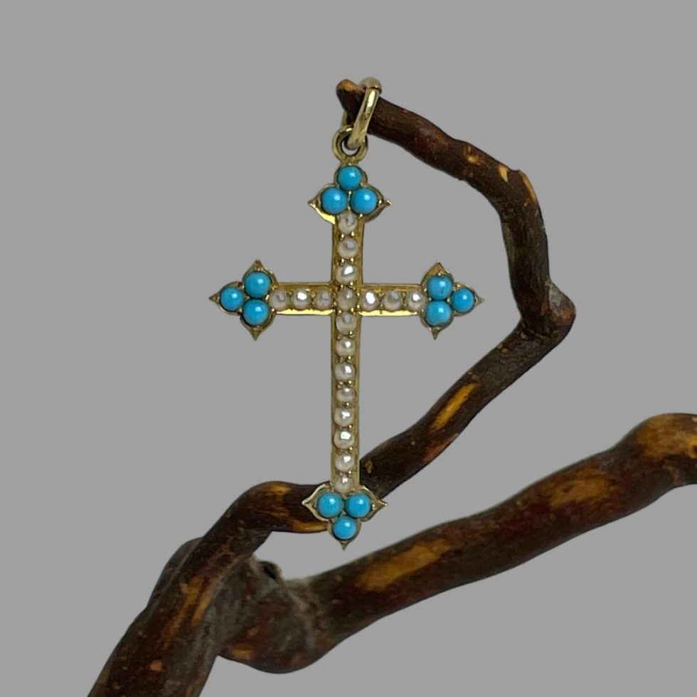 Main photo of antique 18 karat gold cross displayed hanging from a twig of natural cherry wood. The cross is forward facing. The 4 tips of the cross has 3 pointed tips housing 3 small beautiful turquoise blue Sleeping Beauty stones. One stone to pointed tip and 2 immediately behind. 18 seed pearls adorn the rest of the cross. A very pretty piece c1880.