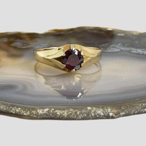 Main photo of gold garnet signet ring seen from an eye level and displayed on a slice of natural agate with the ring front forward facing. The round cut garnet is held in place with 6