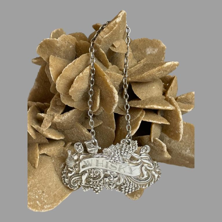 Main photo of silver whisky decanter label displayed hooked and hanging from a rock of natural desert rose. The label area is leaning against the rock at the bottom while the chain is hooked over the top.