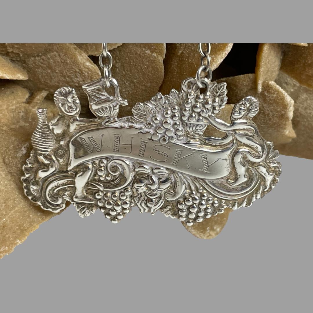 Close up photo of the label area of silver whisky decanter label. At the centre there is a wavy band with WHISKY engraved on. To the left and write of the ribbon are 2 figures