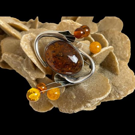 Main photo of sterling silver amber brooch displayed on top of a rock of natural desert sand rose. The generous sized oval central amber is withing a silver frame casing and at the thinner end of the oval at either end, a tube of silver resembling a calla lily flower head curves out and over to opposite side so the heads of the flowers end up at the wide edge of the oval. 3 small balls of amber then protrude from the lily heads. These are mixed egg yolk, butterscotch and cognac amber balls. There is a quite a spacey look about this brooch. It is fun and very funky!